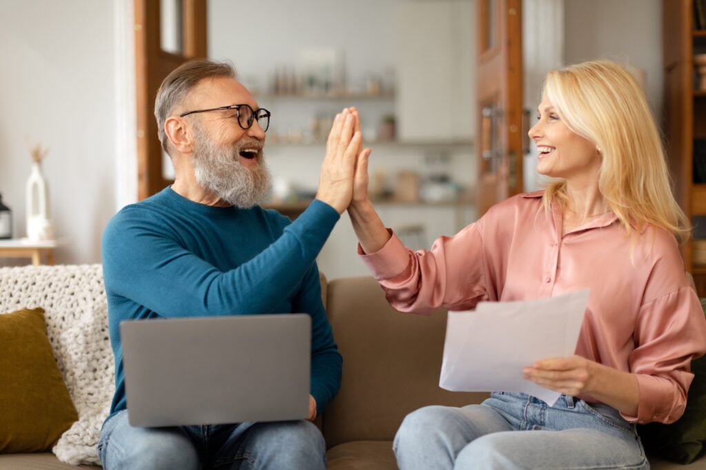 Senior Couple Giving High Five Using Laptop Holding Papers Indoors