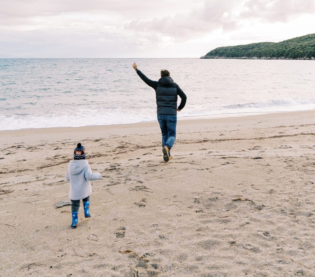 Small child runs after dad with a kite along the beach by the sea. Back view